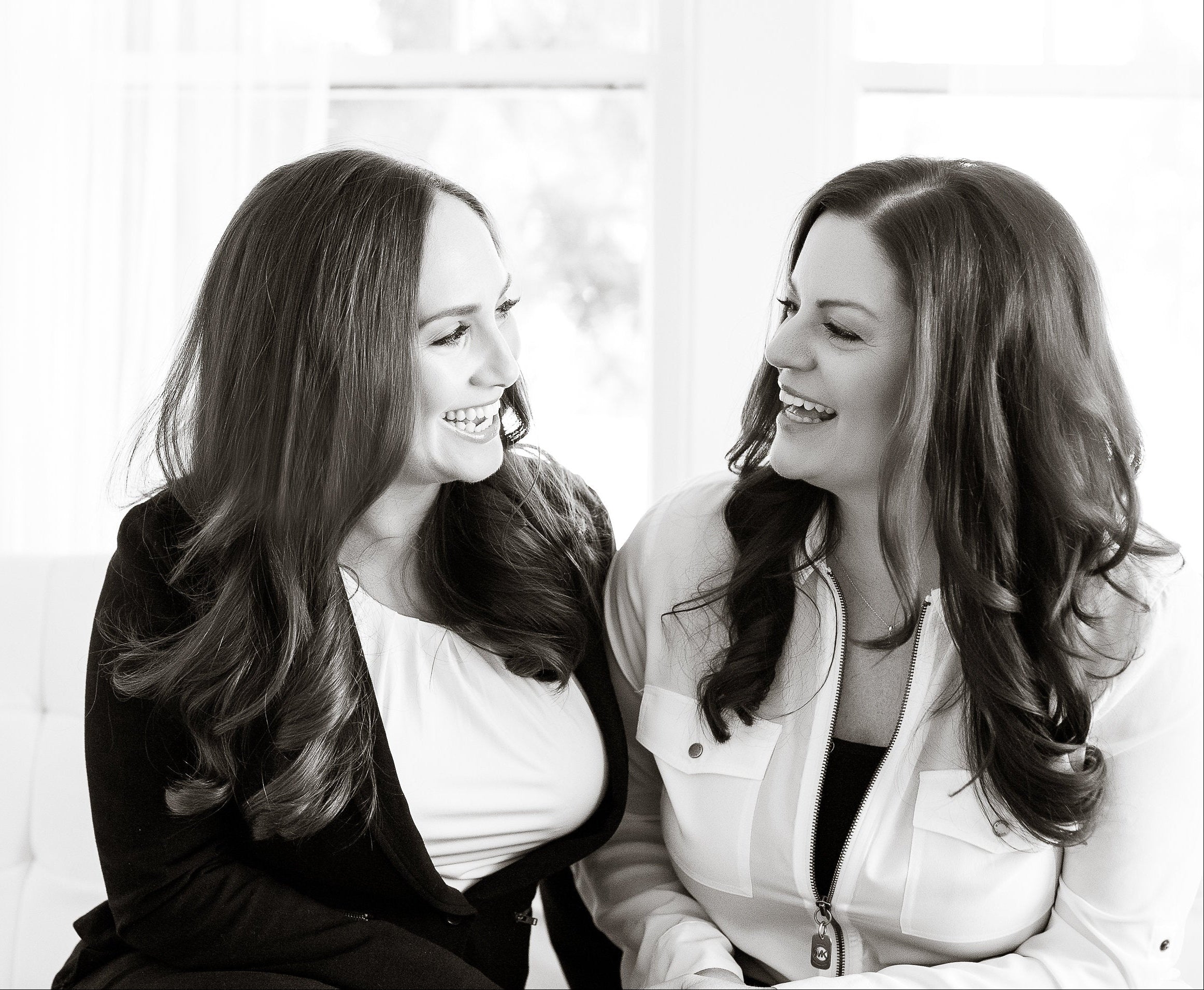 Two women sitting together on a white couch, smiling and looking at each other.