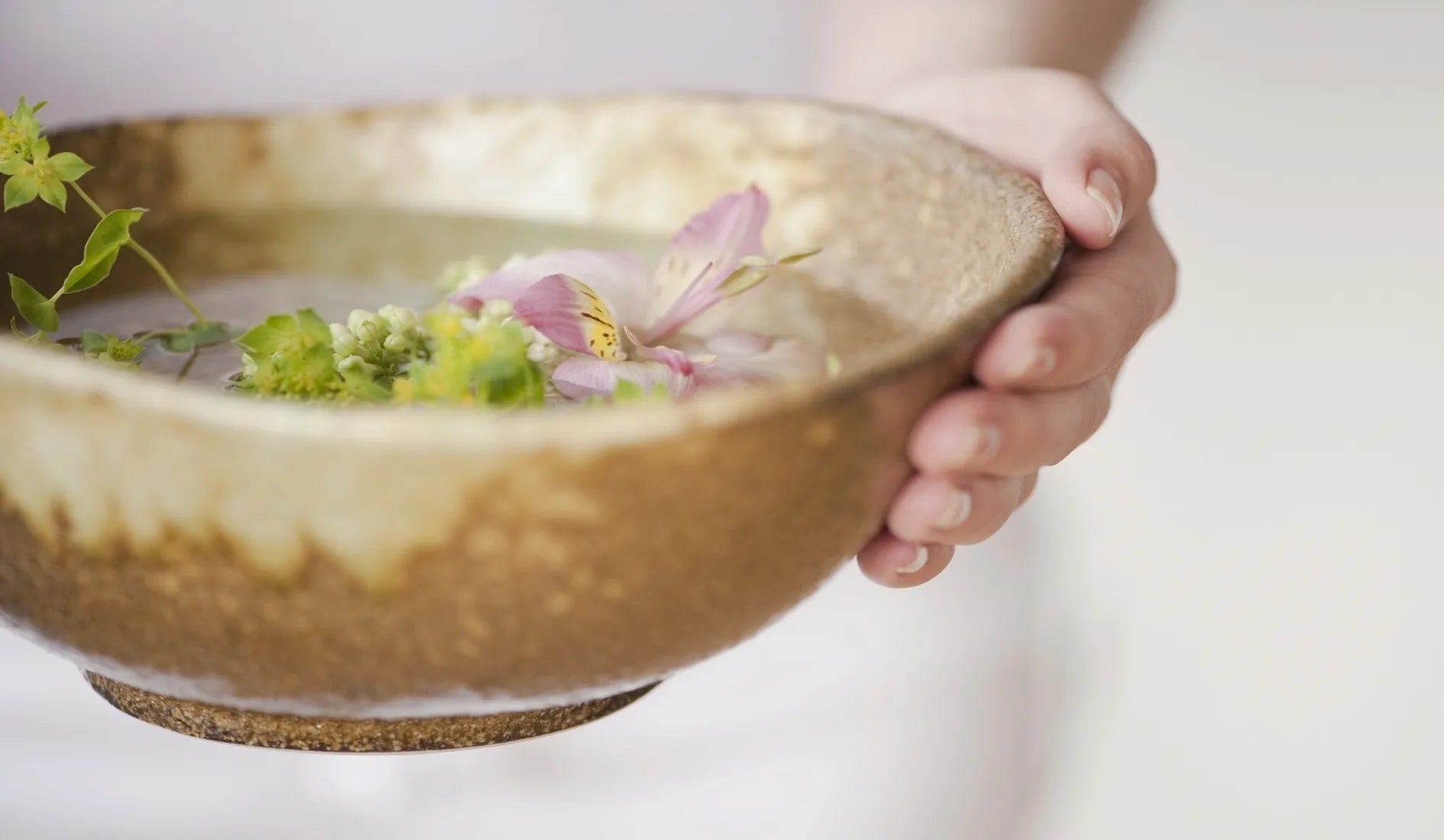 Person holding a bowl with flowers and greenery against a plain background