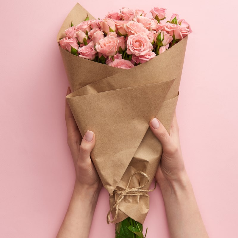 Bouquet of pink roses wrapped in brown paper held by hands on a pink background